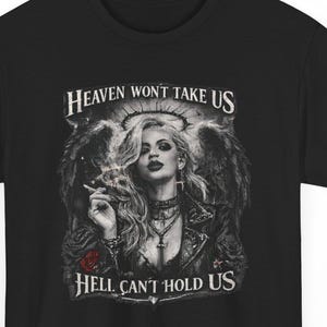 Può includere: T-shirt nera con una grafica dettagliata di una donna con ali d'angelo, un'aureola e una rosa. Il testo "Heaven Won't Take Us" e "Hell Can't Hold Us" è stampato sopra e sotto l'immagine.