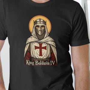 Può includere: T-shirt nera con una grafica di Re Baldovino IV, raffigurato con elmo e veste, con una croce rossa. Il design include un'aureola dorata e il testo "King Baldwin IV" sotto.