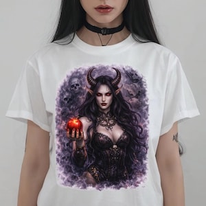 May include: White t-shirt with a fantasy illustration of a horned woman holding a red apple. The artwork has a dark, gothic theme with purple and black accents. The woman has long black hair and is wearing a black corset.