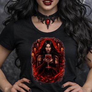 May include: Black t-shirt featuring a gothic illustration of a woman holding a glass of red liquid, surrounded by candles. The woman in the image has long black hair, red eyes, and dark lipstick. A black choker with a red pendant is worn around the neck.