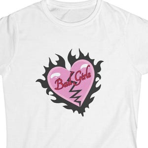 Può includere: T-shirt bianca con un disegno a cuore rosa, una linea spezzata e le parole "Bad Girls" in rosa. Il cuore è incorniciato da un bordo nero a forma di fiamma. La maglietta ha maniche corte e girocollo.