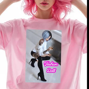 Può includere: Una maglietta rosa chiaro con una grafica di una bambola con i capelli blu, una sigaretta e una bevanda. La grafica è in una cornice rosa con le parole "Fashion Doll". La modella nell'immagine ha i capelli rosa.