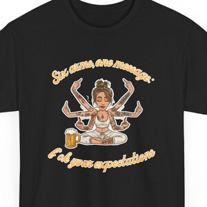 Puede incluir: Camiseta negra con una ilustración de dibujos animados de una mujer con seis brazos en posición de yoga, sosteniendo una jarra de cerveza. El texto dice "Six arms, one message: F*ck your expectations" en amarillo.