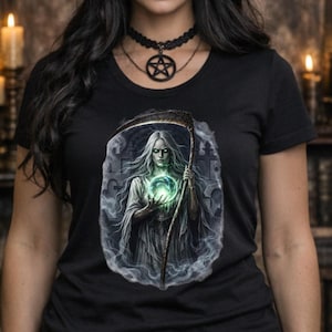 May include: Black t-shirt featuring a detailed graphic of a spectral figure holding a glowing orb and scythe. The person in the image is wearing a black witch hat, choker necklace with a pentagram pendant, and black lipstick. The background includes lit candles.