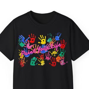 Può includere: T-shirt nera con un design colorato di impronte di mani. La maglietta presenta numerose impronte di mani in vari colori, tra cui rosso, blu, verde e giallo. Le parole "Paint Your Life" sono scritte in rosa sul design.