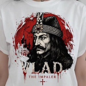 Può includere: Una t-shirt bianca a maniche corte con una stampa grafica di Vlad l'Impalatore con cappello e cappotto scuri su uno sfondo circolare rosso. Il testo recita VLAD THE IMPALER con un simbolo a croce. Un piccolo logo riporta MR DARK-GOTH.