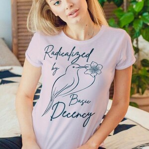 Può includere: T-shirt lilla chiaro con la scritta "Radicalized by Basic Decency" e un disegno di uccello. La persona indossa leggings blu scuro. Lo sfondo include un letto e un divisorio decorativo.