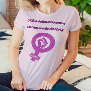 Puede incluir: Camiseta rosa claro con el texto "Well-behaved women seldom make history" y un símbolo feminista morado. La camiseta se lleva con leggings azul oscuro. Una pila de libros está cerca.