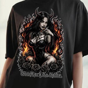 Può includere: T-shirt nera oversize con una grafica di una donna con corna, fiamme e rose. Il testo "Too Dark To Tame" è stampato sotto. La maglietta è indossata da una persona.