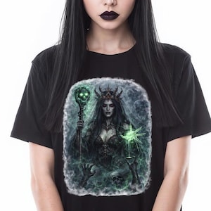 May include: Black t-shirt featuring a fantasy illustration of a dark-haired woman with a crown, holding a staff. The artwork is set against a green and blue smoky background. The woman is wearing a black top and black pants.