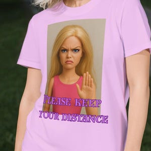 Può includere: Una maglietta lilla con la scritta "PLEASE KEEP YOUR DISTANCE" e una grafica di una bambola. La persona tiene in mano dei pattini a rotelle. La maglietta è a girocollo e maniche corte.