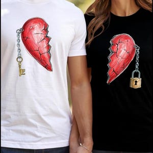 May include: White and black t-shirts with matching broken heart designs. The white shirt features a red heart with a key, while the black shirt has a heart with a padlock. Both shirts are short-sleeved.