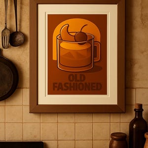 Op de afbeelding: Een ingelijste kunstprint met een illustratie van een "Old Fashioned" cocktail. De afbeelding toont een cocktailglas met een kers en sinaasappelschijf tegen een warme oranje achtergrond. De bruine lijst staat in een keuken.