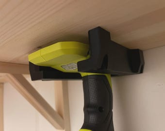 Può includere: Un utensile elettrico nero e giallo è fissato in un supporto nero, montato sotto una mensola di legno. L'impugnatura dell'utensile è nera con un'impugnatura strutturata e il corpo è giallo. Il supporto è fissato sotto la mensola.