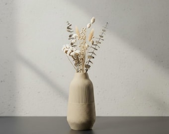 May include: A beige ceramic vase with vertical ribbed detailing on the upper half, holding a bouquet of dried flowers and foliage. The vase sits on a dark surface against a textured white wall, with shadows.