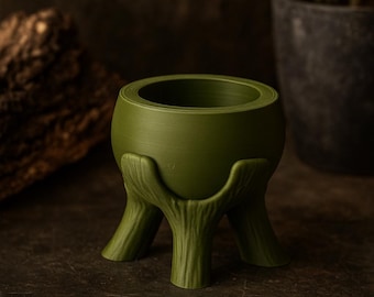 May include: A green planter with a unique tree trunk base. The round pot is a muted green color, supported by three tree trunk-shaped legs. The planter is designed to hold small plants and adds a natural touch to any space.