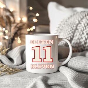 May include: White ceramic mug with the word "ELEVEN" above and below the number "11" in a red and cream color scheme. The mug has a large handle and is sitting on a white surface.
