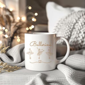 May include: White ceramic mug with the word "Ballerina" in gold script, along with three ballerina figures and a heart. The mug has a large handle and is set against a soft, neutral background.