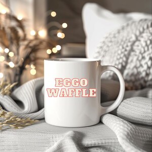 May include: A white ceramic mug with the words "EGGO WAFFLE" in red block letters. The mug has a curved handle and is set against a soft, neutral background with decorative lights.