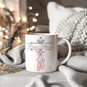 May include: White ceramic mug with the words "Prima Ballerina" in a decorative font, a pink bow, and a pair of pink ballet slippers. The mug has a curved handle and is set against a soft, neutral background.