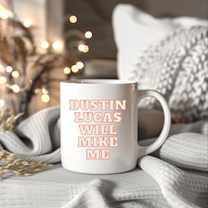 May include: White ceramic mug with the text "DUSTIN LUCAS WILL MIKE ME" in red letters. The mug has a large handle and is sitting on a white surface. The background includes a soft, blurred setting with decorative lights.