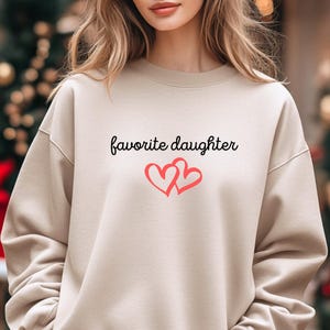 May include: A beige sweatshirt with the words "favorite daughter" in black script and two intertwined red hearts. The sweatshirt has long sleeves and a crew neck.