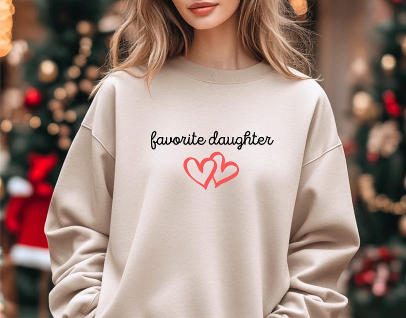 May include: A beige sweatshirt with the words "favorite daughter" in black script and two intertwined red hearts. The sweatshirt has long sleeves and a crew neck.