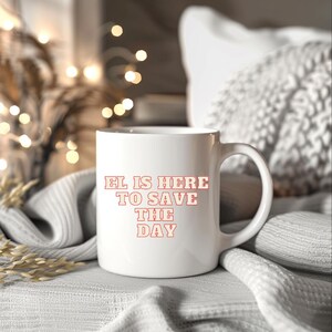 May include: White ceramic mug with the text "EL IS HERE TO SAVE THE DAY" in a red, retro font. The mug is on a white surface with a soft, blurred background.