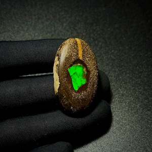 Puede incluir: Una piedra preciosa de ópalo boulder de forma ovalada que presenta una matriz de piedra de hierro texturizada de color marrón oscuro con un único parche de color verde neón vibrante centrado en la piedra, mostrando un tono verde esmeralda.