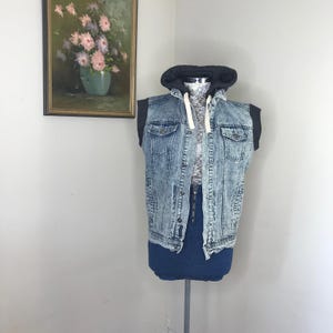 May include: A denim vest with a black hooded sweatshirt attached. The vest has a light blue acid wash design with two chest pockets and button closures. The mannequin is wearing a dark blue denim skirt. The background includes a framed painting of flowers.