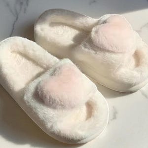 May include: A pair of fluffy white slippers with a light pink heart-shaped accent on the top. The slippers have a soft, plush texture and a thick sole, perfect for indoor wear. The slippers are designed for comfort and relaxation.
