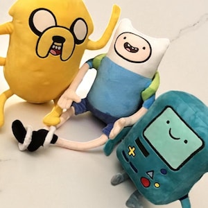 May include: Three plush toys from the animated series Adventure Time. Jake the Dog is yellow, Finn the Human is in blue and white, and BMO is teal. The toys are soft and have embroidered details, perfect for fans of the show.