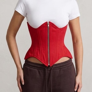 May include: A vibrant red corset featuring a central silver-toned metal zipper closure, structured vertical paneling, and a pointed hemline. The garment is worn over a plain white short-sleeved t-shirt and paired with dark brown drawstring sweatpants.