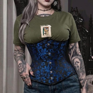 May include: A black and blue corset with a floral pattern. The corset is worn over a green t-shirt with the text "GOVEN UP" and a graphic. The person is wearing blue jeans and has multiple tattoos.