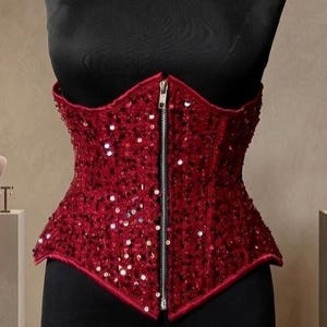 May include: A red sequined corset with a front zipper closure. The corset is displayed on a black mannequin. The garment is a deep red color and covered in small, reflective sequins.