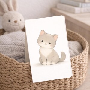Puede incluir: Una tarjeta de felicitación con una ilustración a la acuarela de un gatito gris y blanco. El gato está sentado erguido con la cola enroscada. La tarjeta se muestra en una cesta de mimbre con un conejito de peluche y una manta gris.