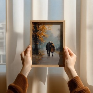 May include: A framed art print depicts an elderly couple walking on a path, embraced. The artwork features a muted color palette with autumn foliage in shades of gold and brown. The frame is made of light-colored wood.