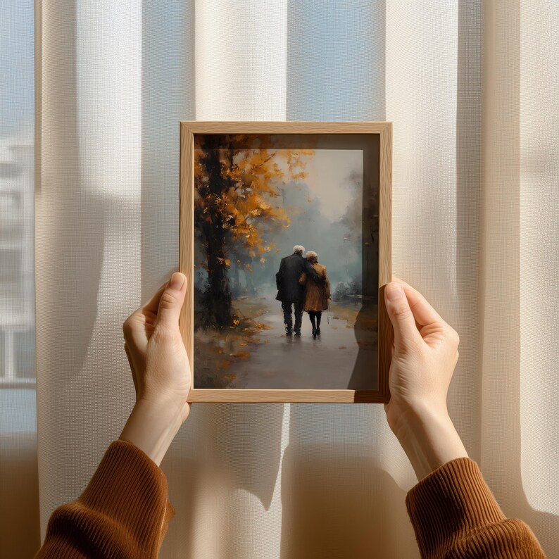 May include: A framed art print depicts an elderly couple walking on a path, embraced. The artwork features a muted color palette with autumn foliage in shades of gold and brown. The frame is made of light-colored wood.
