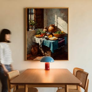 May include: A framed painting depicts a still life with a table set with food and pottery. A blue and red table lamp sits on a wooden table in front of the painting. A person walks by the table.