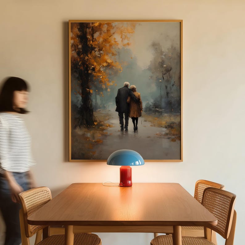 May include: A framed painting depicts an elderly couple walking on a path, with autumn trees in the background. A blue and red table lamp sits on a wooden table. The room also features a wooden chair.