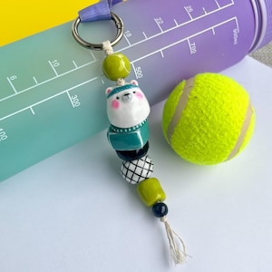 May include: A handmade keychain featuring a ceramic polar bear charm with a teal sweater and a white hat. The keychain has a variety of beads in green, black, and white, and a small tassel. A gradient water bottle and a tennis ball are also in the image.