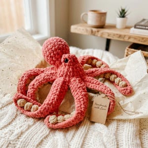 Könnte beinhalten: Handgefertigter Häkel-Oktopus in Korallenrosa mit beigen Akzenten an den Tentakeln. Der Oktopus hat schwarze Augen und ein Etikett mit der Aufschrift "Handmade with Love". Das Plüschtier liegt auf einer cremefarbenen Strickdecke.
