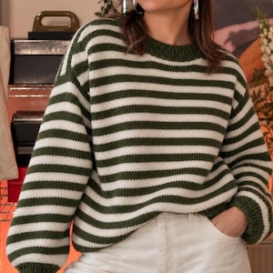 Pode incluir: Uma camisola de malha de manga comprida com riscas horizontais em verde floresta escuro e branco sujo. A peça apresenta um decote redondo, ombros descaídos e punhos canelados nas mangas e na bainha, combinada com calças de ganga brancas.