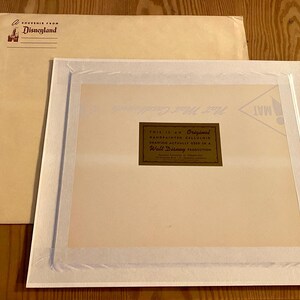 May include: A white matted frame with a gold label that reads "This is an Original Handpainted Cel Animation Drawing Actually Used in a Walt Disney Production". The mat is surrounded by a white border. The frame is on a brown surface.
