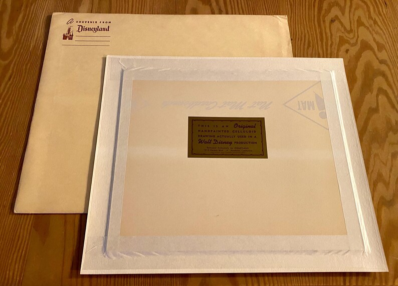 May include: A white matted frame with a gold label that reads "This is an Original Handpainted Cel Animation Drawing Actually Used in a Walt Disney Production". The mat is surrounded by a white border. The frame is on a brown surface.