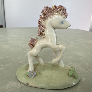 May include: A ceramic unicorn figurine with a white body, pink mane and tail, and gold accents. The unicorn stands on a light green base with a blue flower and green leaves. The figurine is a decorative item.