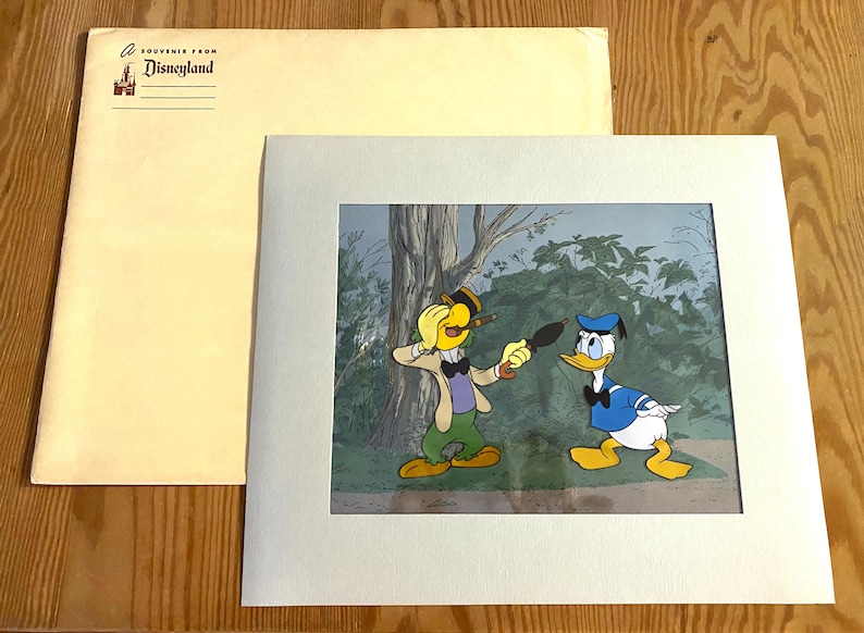 May include: A framed cartoon illustration featuring two characters. One character is a yellow bird wearing a hat and a green vest. The other character is a blue duck wearing a blue hat and a blue bow tie. The illustration is set in a forest scene with green trees and bushes. The frame is white with a light gray mat.