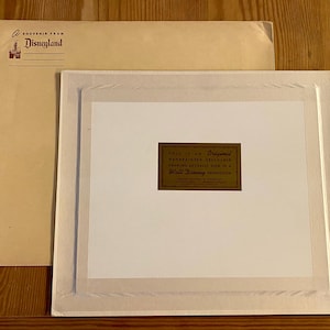 K&ouml;nnte beinhalten: Ein wei&szlig;er Rahmen mit Passepartout und einem goldenen Etikett mit der Aufschrift "This is an Original Handpainted Cellluloid Drawing Actually Used in a Walt Disney Production".