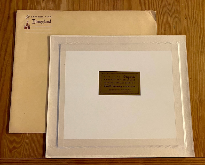 K&ouml;nnte beinhalten: Ein wei&szlig;er Rahmen mit Passepartout und einem goldenen Etikett mit der Aufschrift "This is an Original Handpainted Cellluloid Drawing Actually Used in a Walt Disney Production".