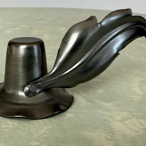 May include: A dark metallic candle holder with a leaf design. The base is a wide, ruffled shape, supporting a cylindrical cup. A stylized leaf curves gracefully from the side, adding a decorative touch. The overall aesthetic is modern and elegant.